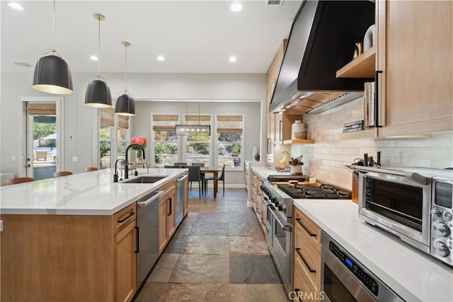 kitchen with appliances with stainless steel finishes, a sink, stone tile floors, wall chimney range hood, and an island with sink
