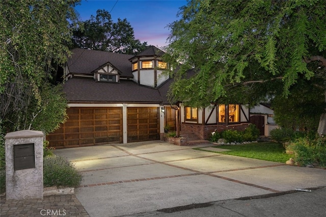 tudor-style house with driveway, an attached garage, brick siding, and roof with shingles