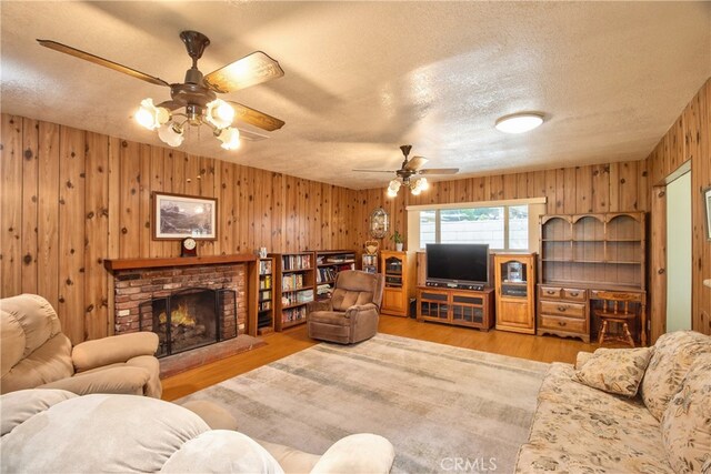 living room with ceiling fan, wooden walls, a textured ceiling, a brick fireplace, and wood finished floors