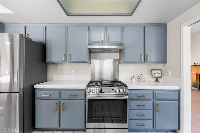 kitchen featuring stainless steel appliances, under cabinet range hood, blue cabinetry, and tile countertops