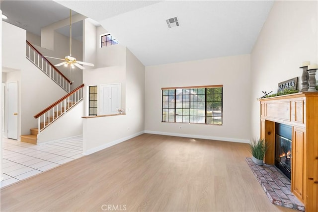 unfurnished living room featuring light wood finished floors, a glass covered fireplace, a ceiling fan, stairs, and high vaulted ceiling