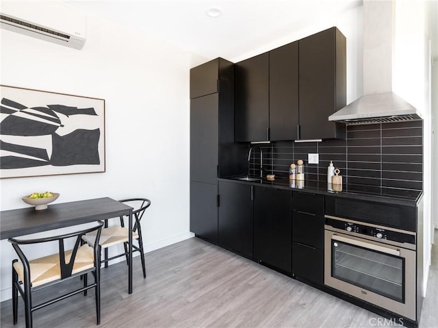 1375 Fortuna Ave, Landers, CA 92285 - kitchen featuring stainless steel oven, wall chimney range hood, light wood finished floors, tasteful backsplash, and a wall mounted air conditioner