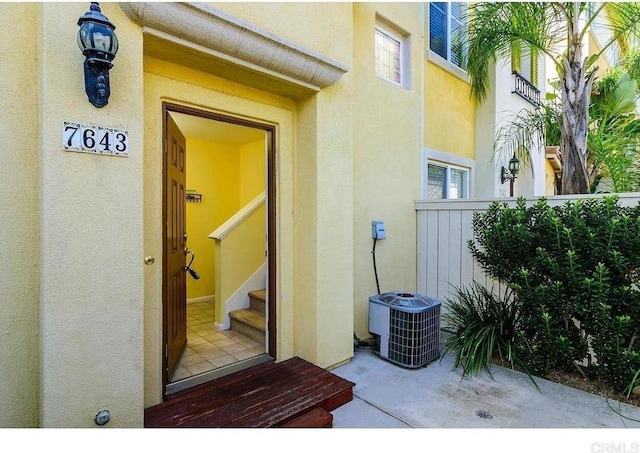 view of exterior entry featuring cooling unit, stucco siding, and fence