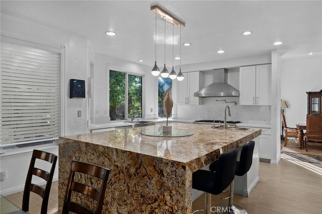 kitchen with wall chimney range hood, an island with sink, a kitchen bar, white cabinets, and recessed lighting