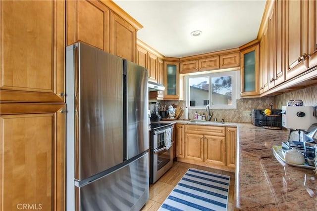 kitchen featuring appliances with stainless steel finishes, a sink, under cabinet range hood, backsplash, and glass insert cabinets