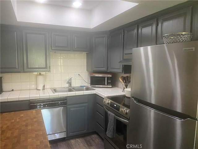 kitchen featuring stainless steel appliances, tile countertops, a sink, decorative backsplash, and gray cabinets