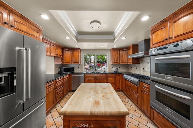 kitchen with stainless steel appliances, butcher block countertops, a raised ceiling, wall chimney exhaust hood, and recessed lighting