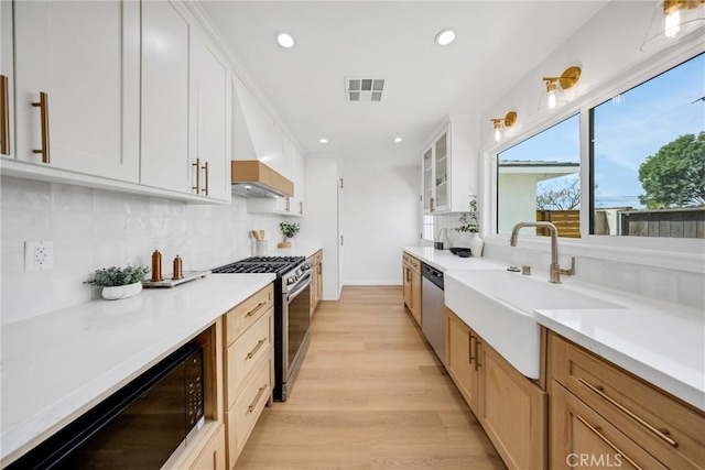kitchen featuring stainless steel appliances, a sink, light wood finished floors, glass insert cabinets, and backsplash