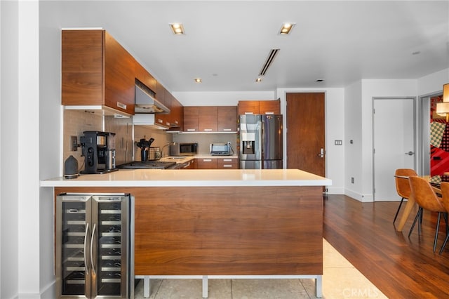 kitchen featuring stainless steel appliances, wine cooler, brown cabinets, under cabinet range hood, and light countertops