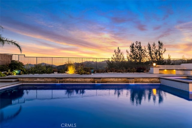pool at dusk featuring a pool with connected hot tub and fence