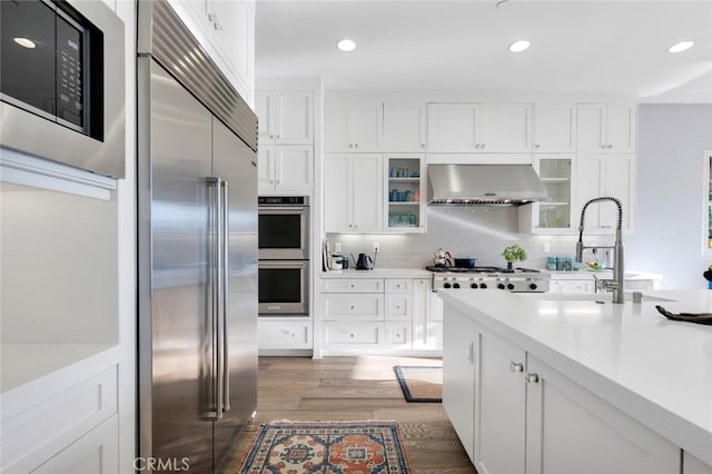kitchen with built in appliances, wall chimney exhaust hood, wood finished floors, light countertops, and white cabinetry