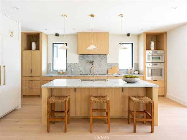 kitchen featuring stainless steel double oven, backsplash, light wood-style floors, an island with sink, and light stone counters