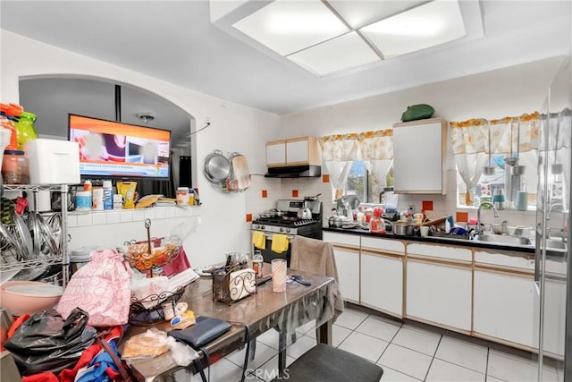 kitchen featuring stainless steel gas range oven, light tile patterned flooring, under cabinet range hood, white cabinetry, and arched walkways