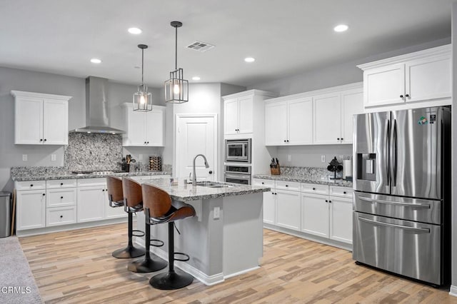 kitchen with appliances with stainless steel finishes, wall chimney range hood, a sink, light wood-type flooring, and white cabinetry