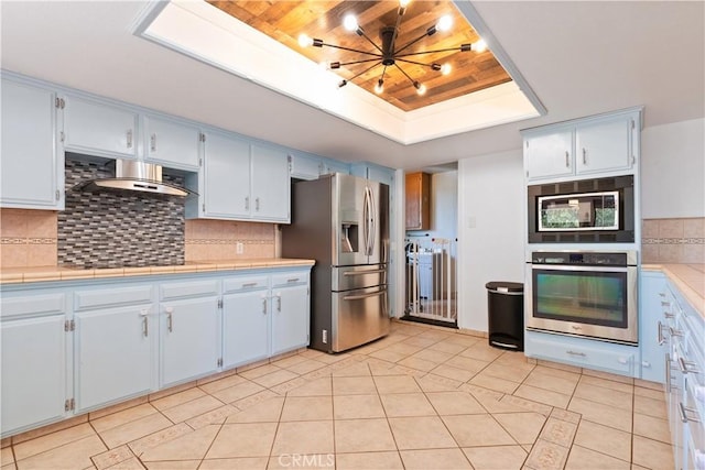 kitchen featuring appliances with stainless steel finishes, a raised ceiling, wall chimney exhaust hood, light tile patterned floors, and wooden ceiling