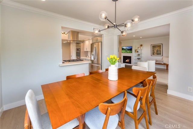 dining space featuring crown molding, baseboards, a warm lit fireplace, a chandelier, and light wood-style flooring
