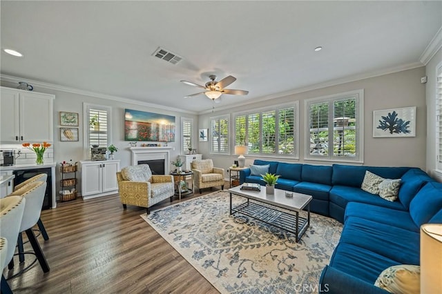 living area with ornamental molding, healthy amount of natural light, dark wood-style floors, ceiling fan, and a fireplace