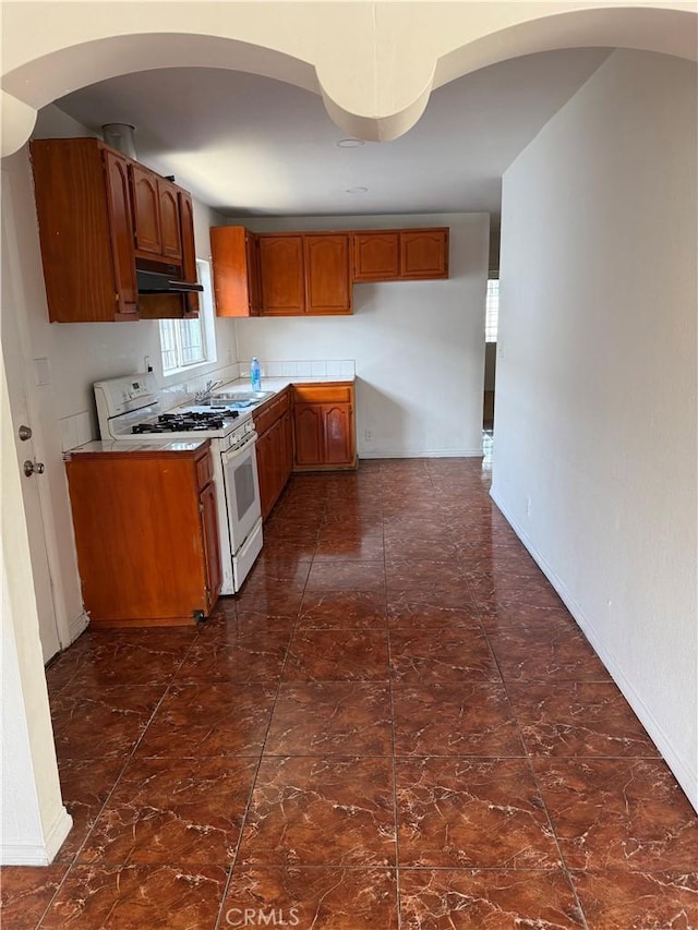 8711 Beach St, Los Angeles, CA 90002 - kitchen featuring white gas range, under cabinet range hood, brown cabinets, light countertops, and arched walkways
