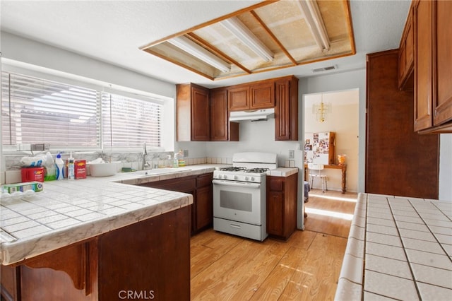 kitchen with white gas range oven, under cabinet range hood, tile counters, and a sink