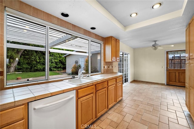 kitchen featuring dishwasher, a sink, tile counters, a ceiling fan, and a tray ceiling