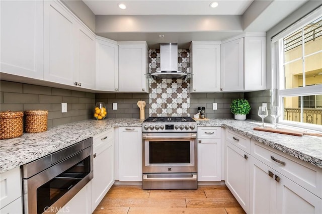 kitchen with appliances with stainless steel finishes, wall chimney range hood, white cabinetry, light wood-type flooring, and decorative backsplash