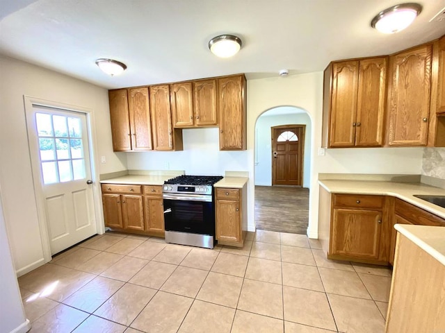 kitchen with stainless steel gas range oven, light tile patterned floors, arched walkways, light countertops, and brown cabinetry