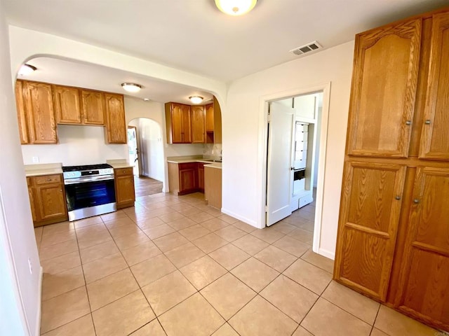 kitchen with stainless steel range with gas cooktop, arched walkways, light countertops, brown cabinets, and light tile patterned floors