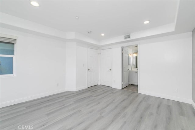 unfurnished bedroom with light wood-type flooring, a tray ceiling, recessed lighting, baseboards, and connected bathroom