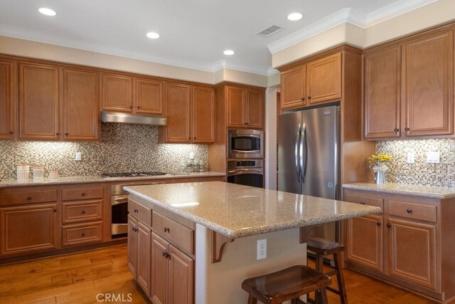 kitchen with appliances with stainless steel finishes, under cabinet range hood, a center island, a breakfast bar, and brown cabinets
