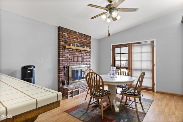 dining area featuring vaulted ceiling, light wood-type flooring, a ceiling fan, a brick fireplace, and baseboards