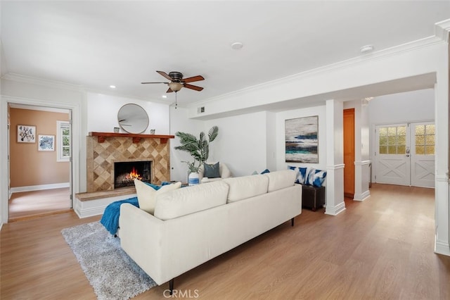 living area featuring ornamental molding, a ceiling fan, light wood-style floors, a tiled fireplace, and baseboards