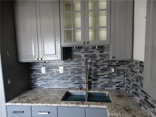 kitchen featuring a sink, backsplash, glass insert cabinets, and light stone countertops