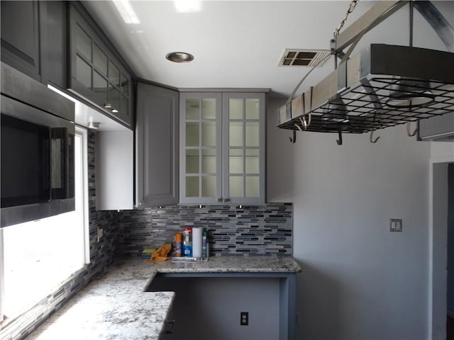 kitchen featuring gray cabinetry, decorative backsplash, glass insert cabinets, and stainless steel microwave