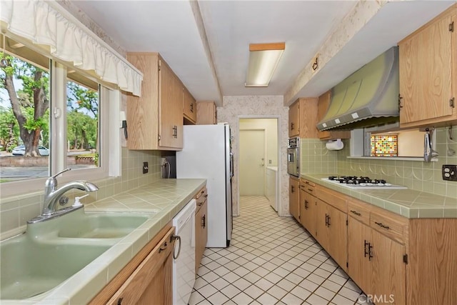 kitchen featuring tile countertops, wall chimney range hood, white appliances, and a sink