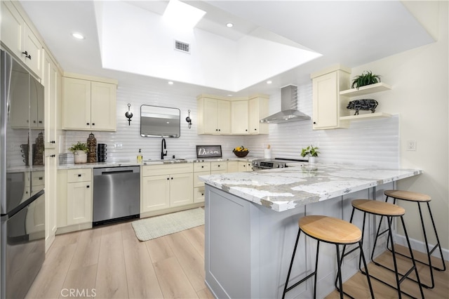 kitchen featuring appliances with stainless steel finishes, wall chimney exhaust hood, cream cabinetry, a peninsula, and recessed lighting