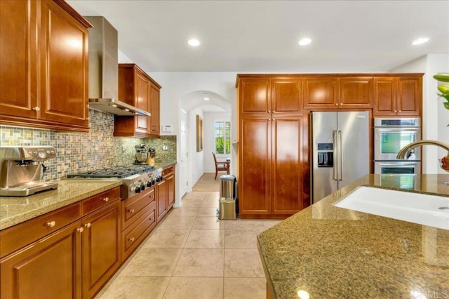 kitchen featuring appliances with stainless steel finishes, wall chimney range hood, arched walkways, tasteful backsplash, and stone counters