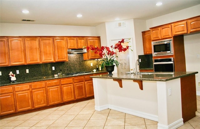kitchen featuring appliances with stainless steel finishes, a sink, dark stone counters, light tile patterned flooring, and under cabinet range hood