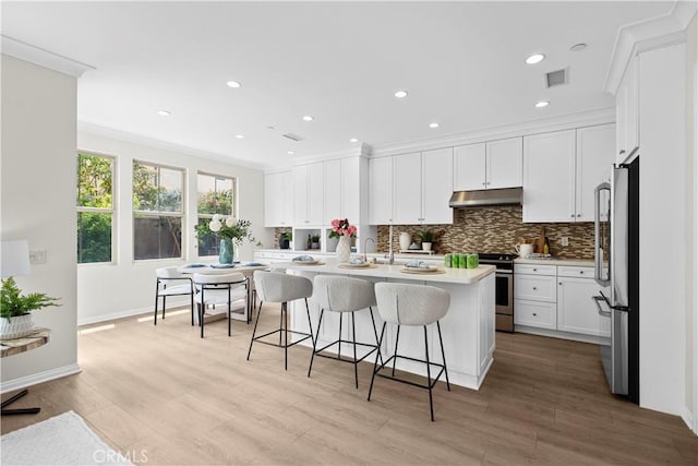 kitchen featuring appliances with stainless steel finishes, under cabinet range hood, crown molding, backsplash, and a breakfast bar area