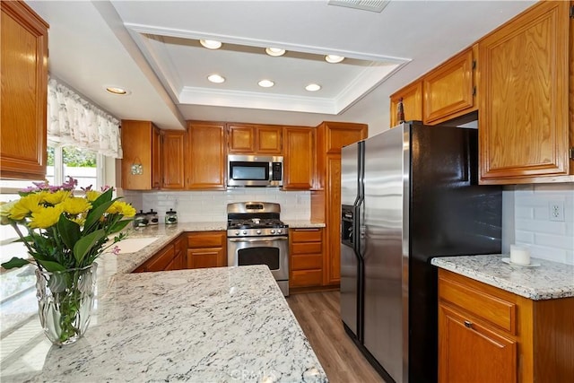 kitchen featuring stainless steel appliances, a tray ceiling, wood finished floors, brown cabinetry, and recessed lighting
