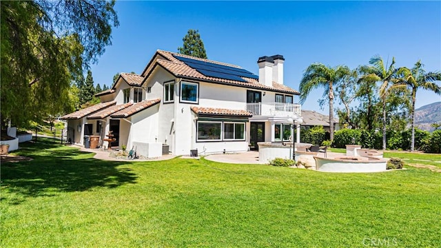 rear view of house featuring a balcony, a chimney, solar panels, a patio, and a lawn