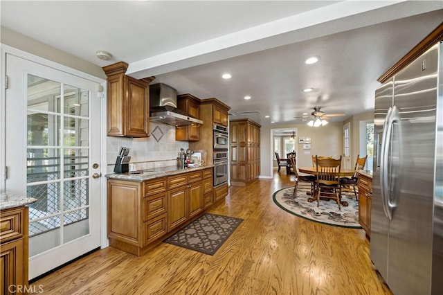 kitchen featuring appliances with stainless steel finishes, wall chimney exhaust hood, brown cabinetry, light wood-style floors, and ceiling fan