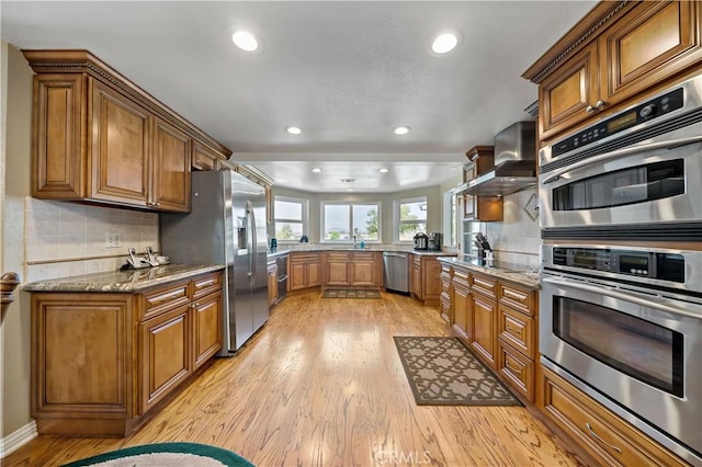 kitchen featuring brown cabinetry, light wood-type flooring, stainless steel appliances, wall chimney range hood, and recessed lighting