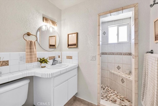 full bath with a textured wall, toilet, a tile shower, vanity, and backsplash