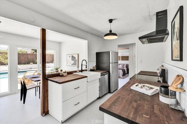 kitchen featuring butcher block countertops, wall chimney range hood, a sink, stainless steel appliances, and white cabinets