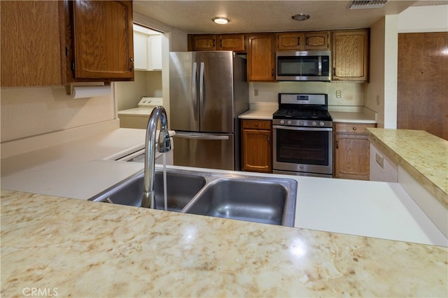 kitchen with stainless steel appliances, light countertops, brown cabinetry, a sink, and washer / dryer
