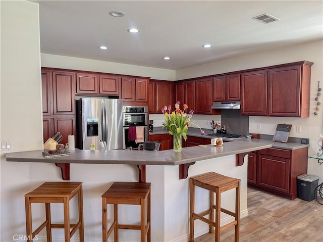 kitchen with appliances with stainless steel finishes, under cabinet range hood, a breakfast bar area, light wood finished floors, and a peninsula