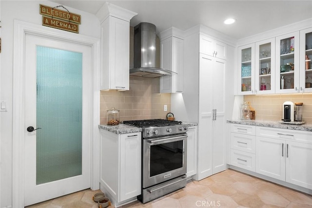 kitchen featuring stainless steel range with gas cooktop, wall chimney range hood, decorative backsplash, white cabinetry, and glass insert cabinets