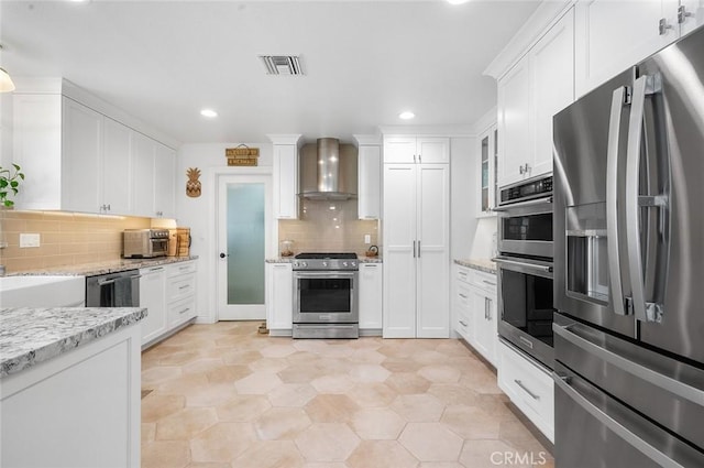 kitchen with stainless steel appliances, wall chimney range hood, white cabinets, tasteful backsplash, and light stone counters