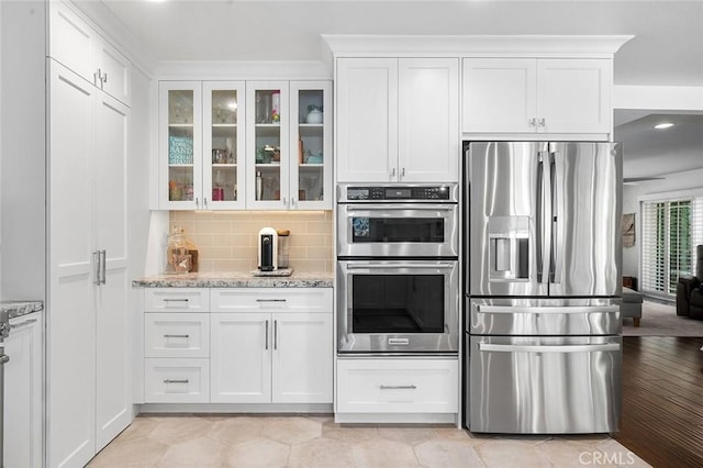 kitchen with stainless steel appliances, glass insert cabinets, white cabinets, decorative backsplash, and light stone counters