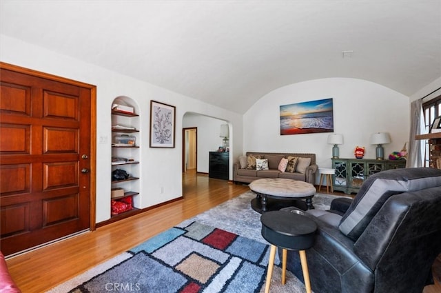 living room with built in shelves, arched walkways, wood finished floors, vaulted ceiling, and baseboards
